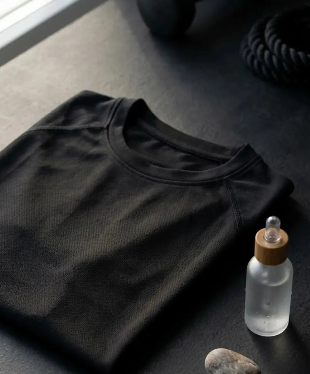 An angled, close-up shot of a neatly folded black, mesh-patterned athletic t-shirt on a dark, textured surface. Next to it are a clear glass serum bottle with a bamboo top, two smooth gray stones, a dumbbell, and black coiled rope, near a window.