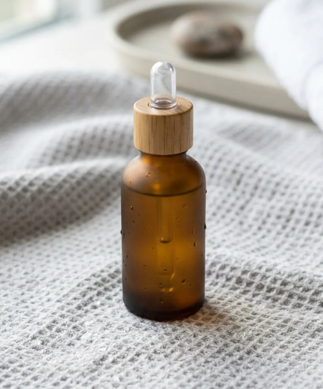 A vertical shot of a minimalist frosted brown glass dropper bottle with a wooden cap, sitting on a light gray waffle knit towel. The bottle has water droplets on it. Soft light comes from a window in the blurred background, creating a clean aesthetic.